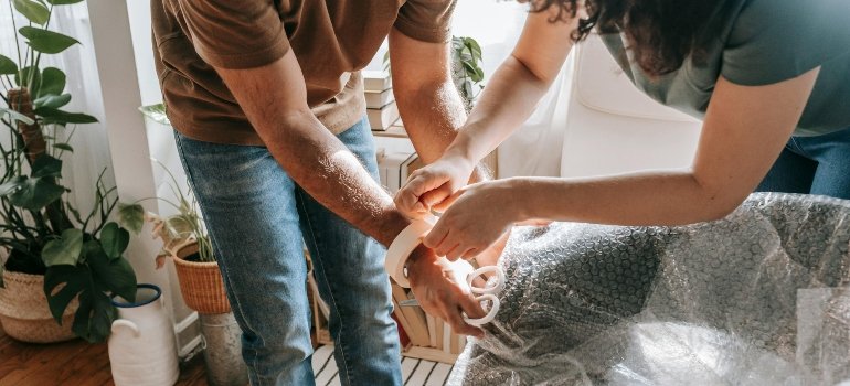 Two people carefully wrapping a chair in plastic to protect it from scratches and dirt during transport, showing how to pack furniture when moving.