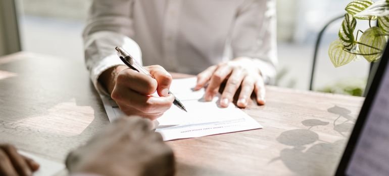 a hand signing a lease document at a desk with a laptop nearby
